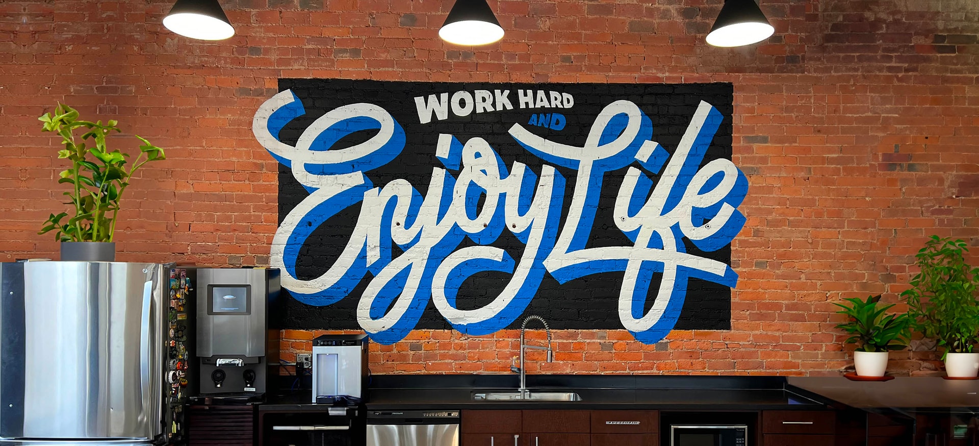 QSC's Ft. Wayne office kitchenette with exposed brick wall featuring large mural of company value “Work hard and enjoy life” in blue and white text.