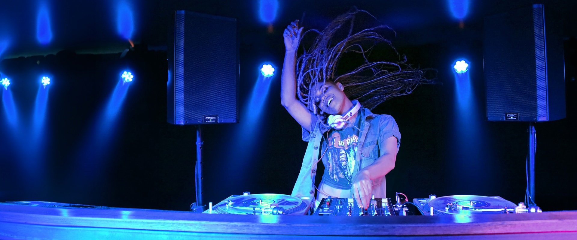 Energetic DJ with dreadlocks performing under vibrant blue stage lighting.