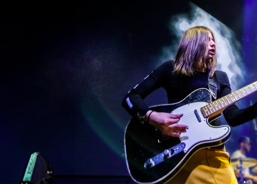 Energetic musician with long hair playing electric guitar on stage with dramatic lighting.