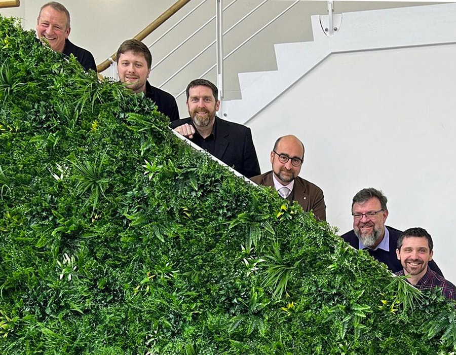 Team of smiling QSC employees posed on stairs, partially obscured by leafy plants in the foreground.